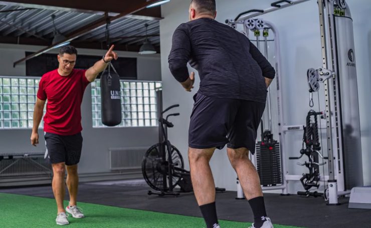 Back view of muscular man in sportswear doing exercises with trainer in modern fitness club